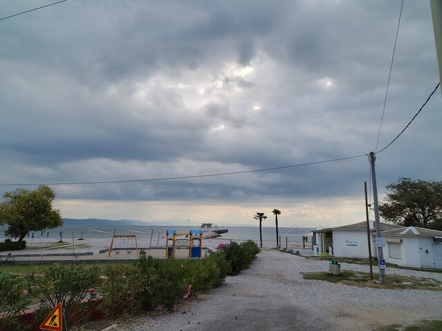 Strand von Monfalcone unter dunklen Wolken, mit Spielplatz, Palmen und einem Steg ins Meer