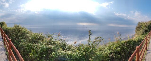 Panorama über das Meer vor Monfalcone, Sonnenlicht bricht durch die Wolken, im Vordergrund Büsche und ein rotes Geländer am Radweg