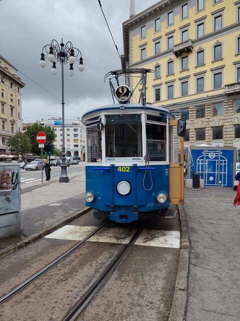 Blau-weiße historische Straßenbahn mit der Nummer 2 und der Aufschrift Trieste–Opicina an der Haltestelle vor gelben Stadthäusern in Triest