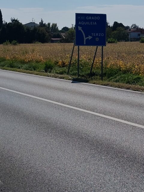 Blaues Straßenschild Richtung Grado und Aquileia an einer Landstraße neben einem Maisfeld